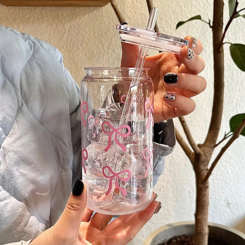 A hand holds a clear glass tumbler filled with water and ice, featuring a pink bow design, alongside a matching pink bow stra