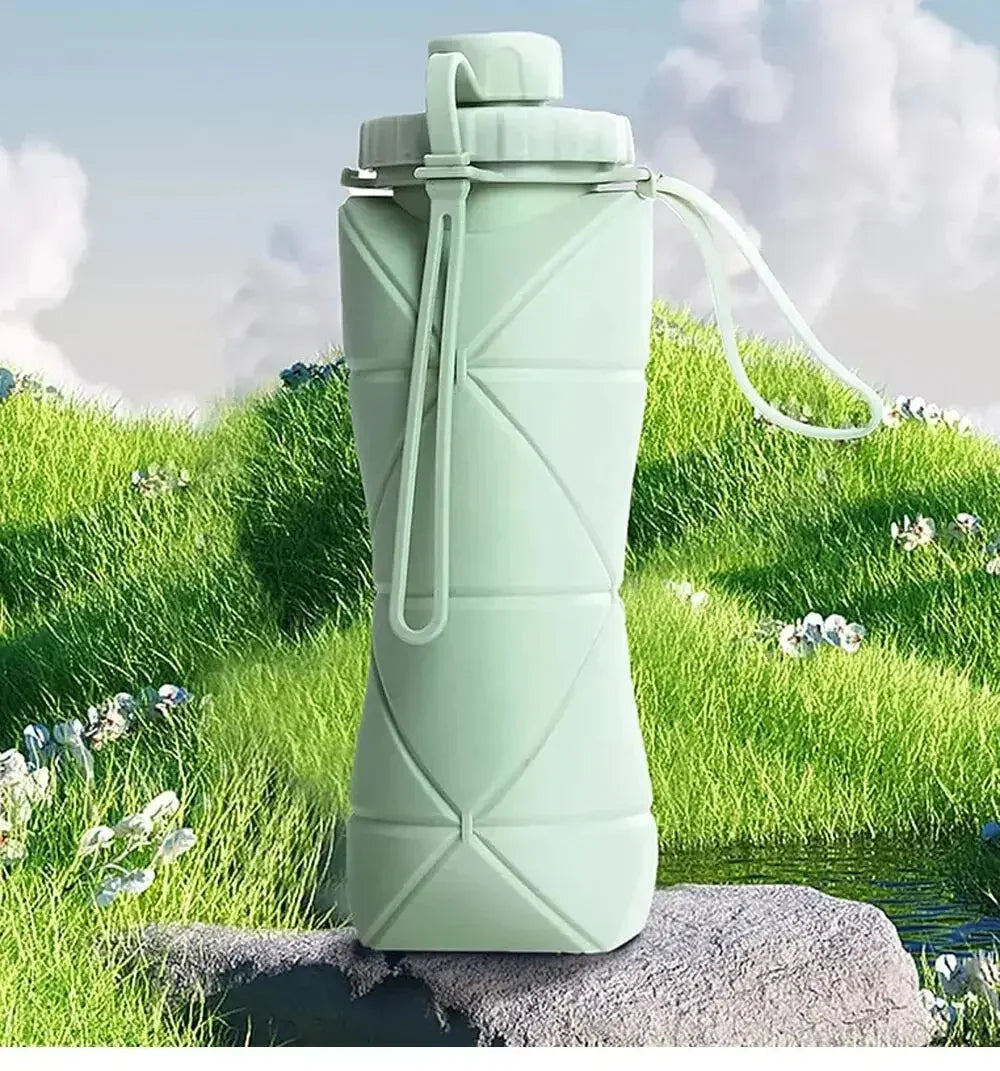 Botella de agua de silicona verde claro y plegable, apoyada sobre una roca con césped y vegetación frondosa.