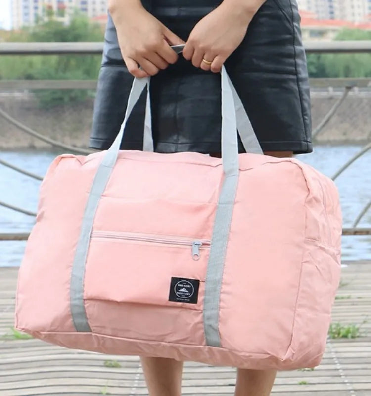 A person is holding a light pink foldable travel duffel bag with gray straps outdoors.