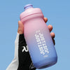 A hand holds a pink and purple squeezable cycling water bottle against a light blue sky.
