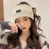A woman models a beige knitted headband while wearing a patterned sweater.