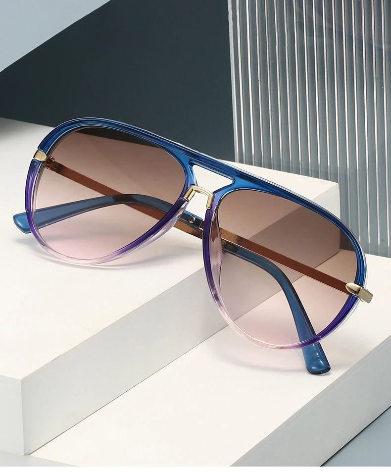 A pair of retro pilot sunglasses with gradient purple and pink lenses resting on a white surface.