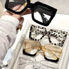 A hand holds a pair of black oversized rectangular reading glasses, with three other pairs displayed in a white tray.
