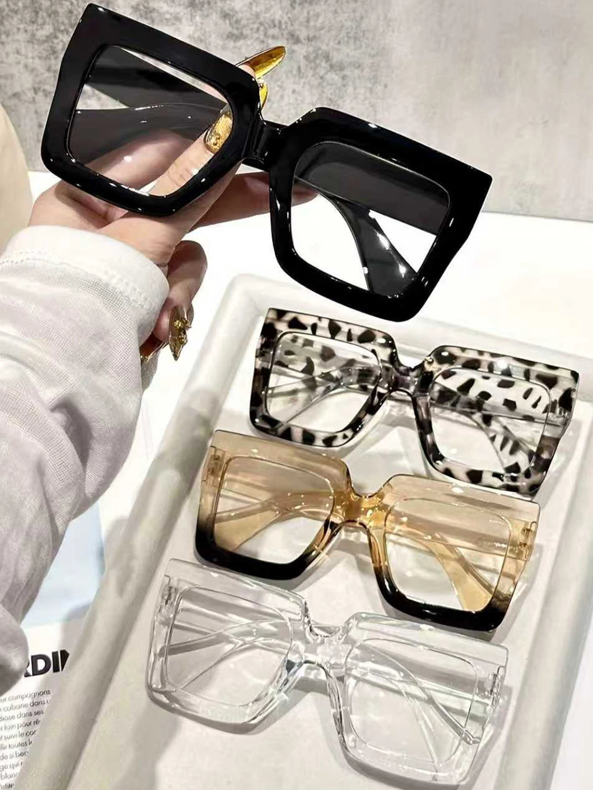 A hand holds a pair of black oversized rectangular reading glasses, with three other pairs displayed in a white tray.