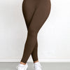 A woman wearing brown high-waisted sports leggings and white sneakers standing against a white wall.
