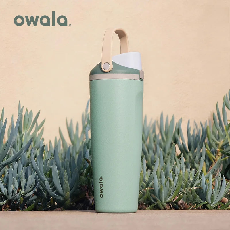 A light green Owala insulated stainless steel travel mug standing in front of blurred succulent plants.