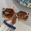 A hand holds a pair of geometric rhinestone fashion sunglasses with brown lenses and silver frames.