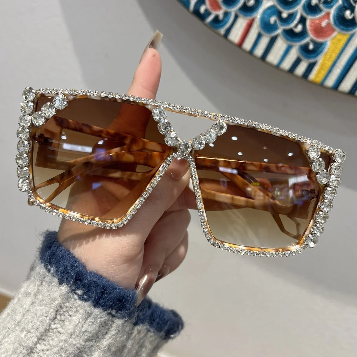 A hand holds a pair of geometric rhinestone fashion sunglasses with brown lenses and silver frames.