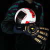 A hand wearing black goalkeeper gloves holds a white and red soccer ball against a dark background.