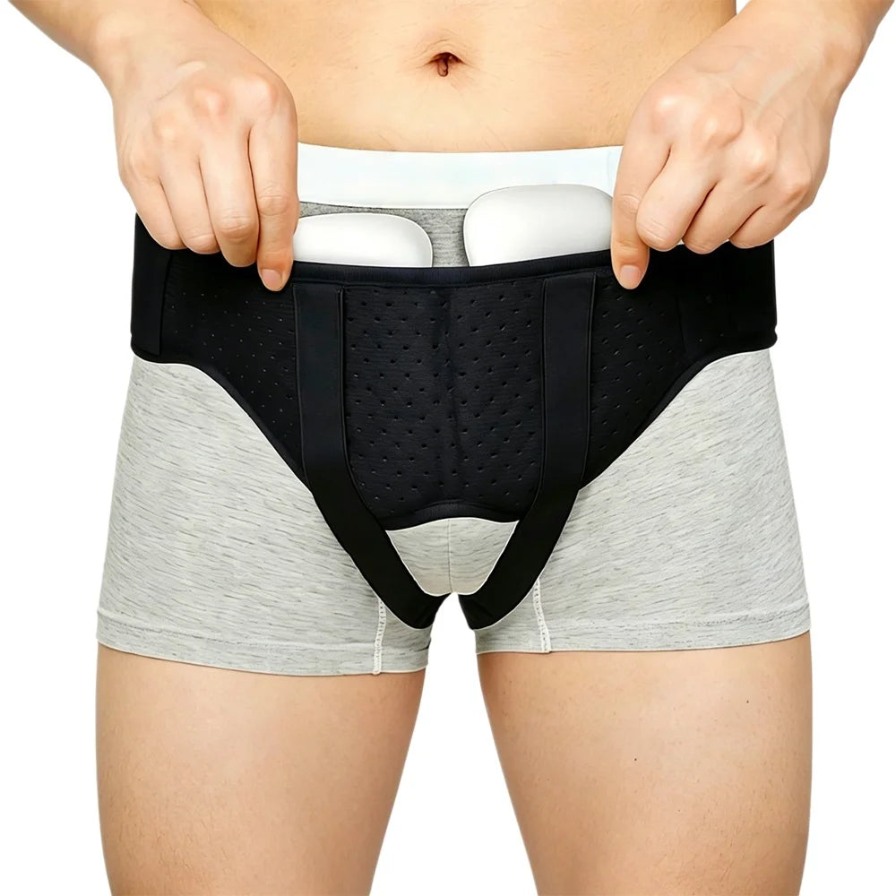 A person is holding an adjustable hernia belt with dual compression pads over light gray shorts.
