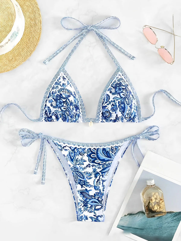 A blue and white triangle halter bikini set laid out on a white marble surface with a straw hat and a small glass jar.