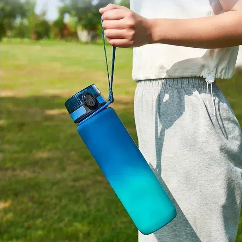 A hand holds a bright blue sports water bottle with a teal cap outdoors on a grassy field.