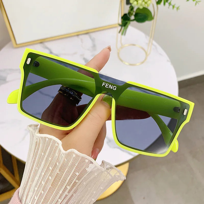 A hand holds a pair of bright lime green oversized square sunglasses with gold accents.