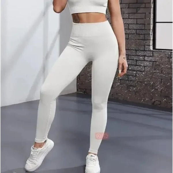 Una mujer vistiendo leggings de yoga de punto sin costuras blancos, de pie en interiores contra una pared de ladrillos.