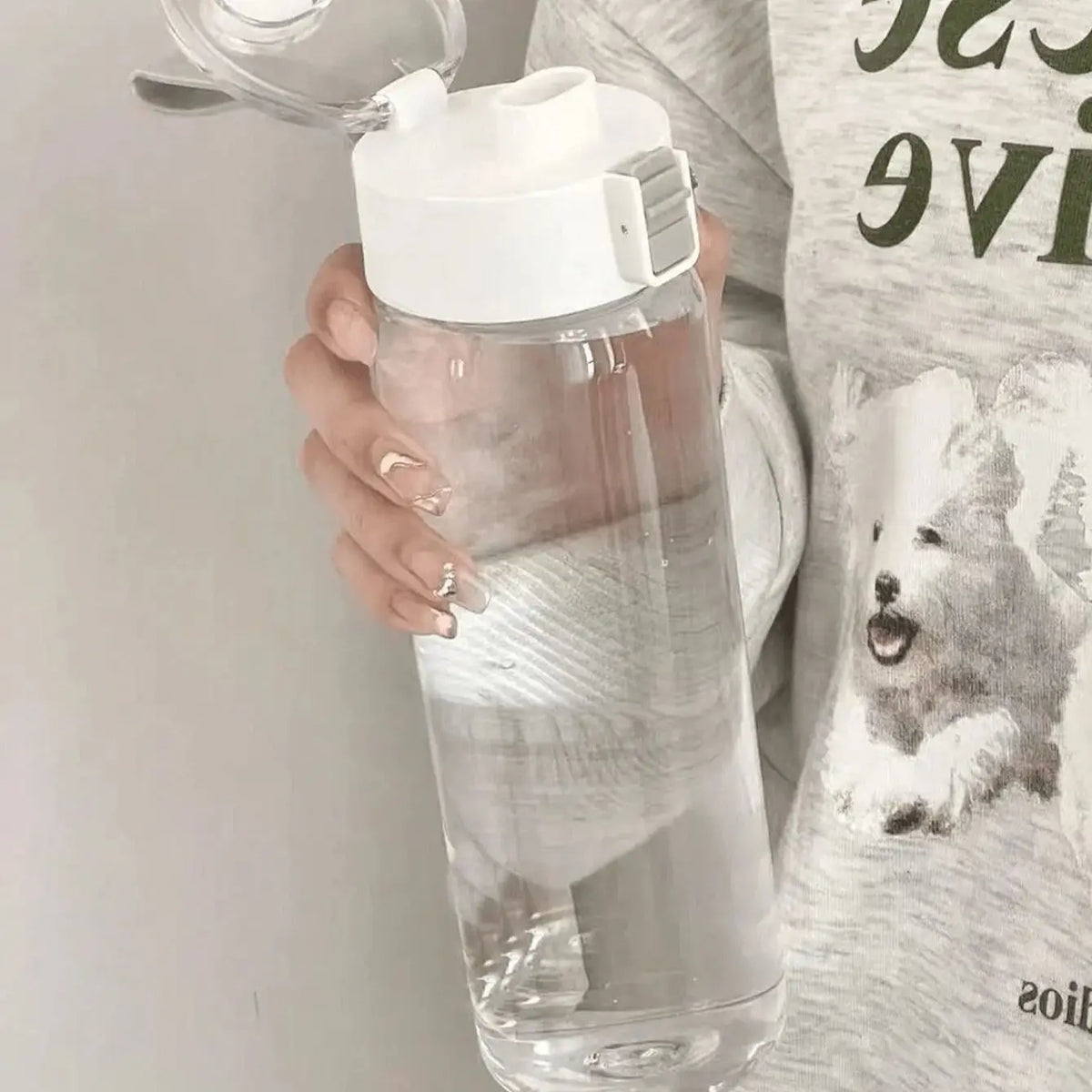 A hand holds a large capacity transparent sports water bottle with a white cap against a light background.