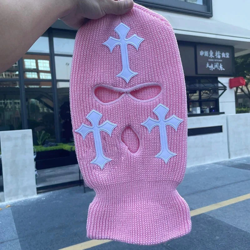 A pink knitted balaclava beanie hat featuring white cross embroidery.