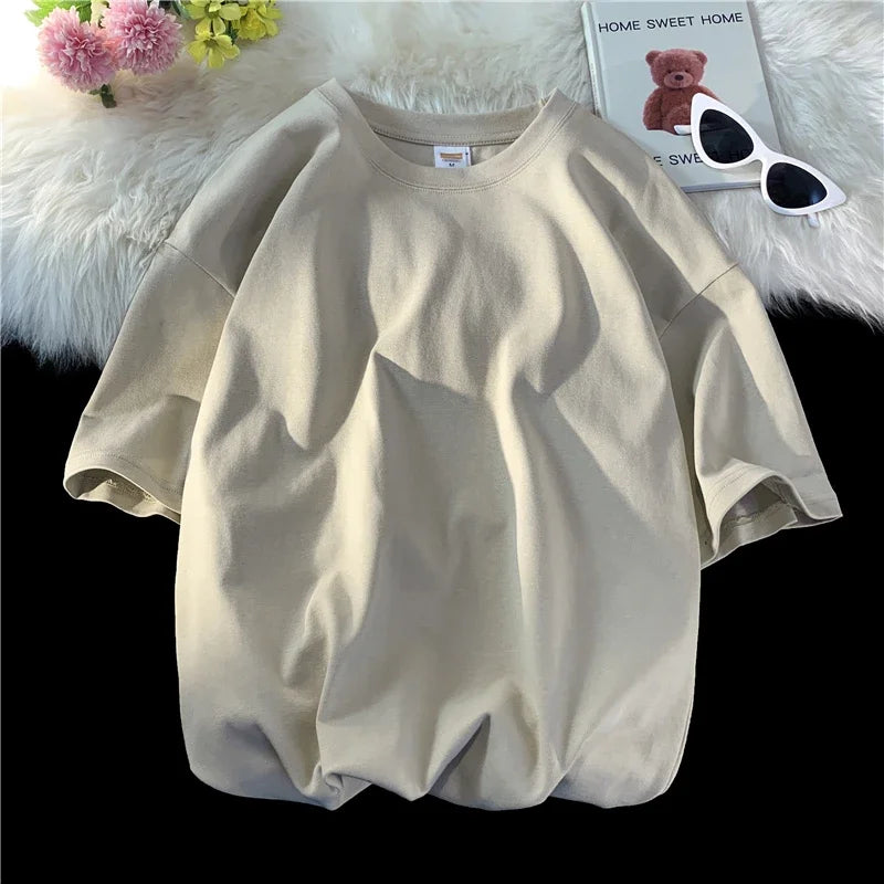 Beige, überdimensioniertes Baumwoll-T-Shirt einer Frau auf weißem Plüsch neben einem Teddybär und Sonnenbrille.