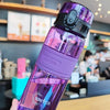 A hand holds a purple glass water bottle with a black cap and tea infuser in a modern cafe setting.