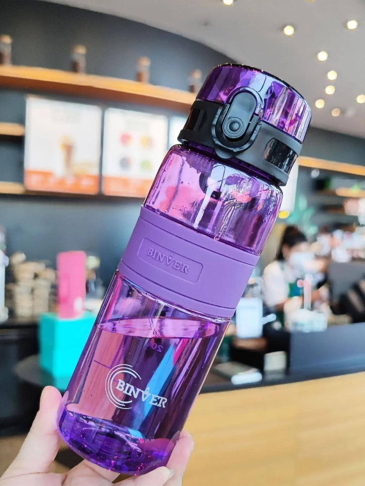 A hand holds a purple glass water bottle with a black cap and tea infuser in a modern cafe setting.