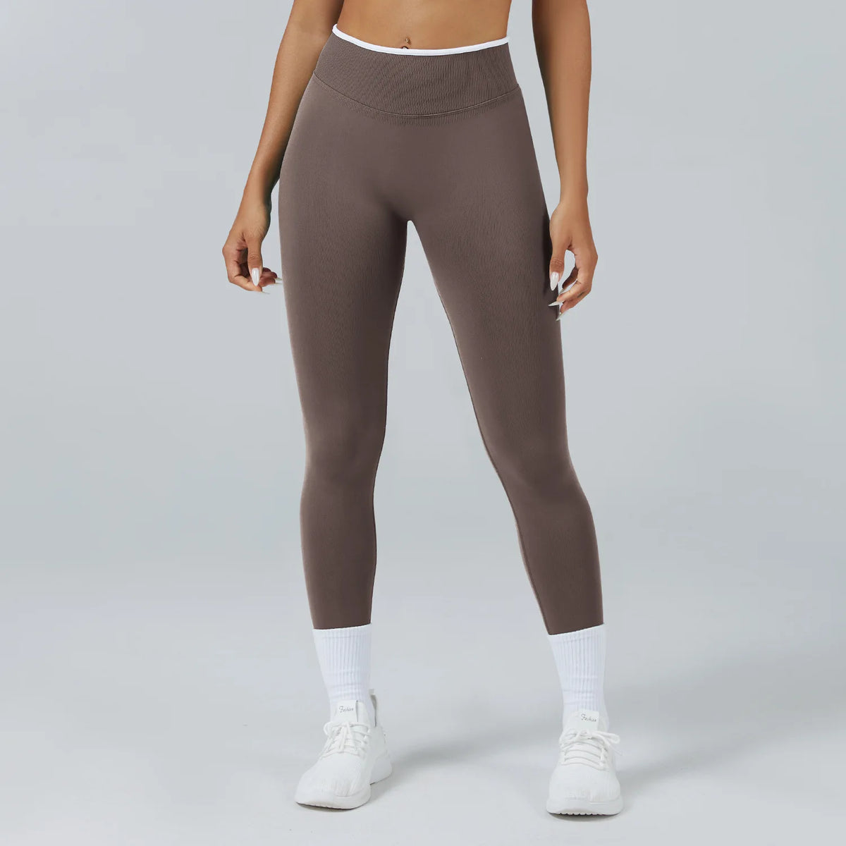 A woman wearing high-waist, full-length, dark brown yoga leggings and white socks, standing against a light gray background.