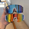 A hand holds a pair of mirrored cycling sunglasses with blue and rainbow colored lenses.
