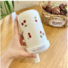 Un tumbler isotherme blanc avec un motif cerise est tenu par une main, placé à côté d'un panier de plantes.