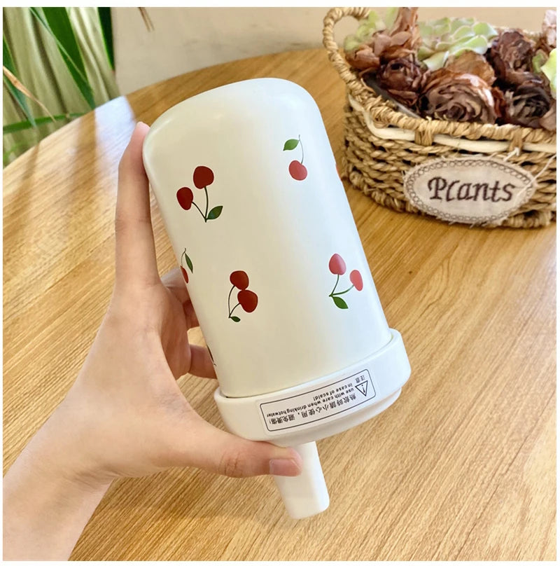 Un tumbler isotherme blanc avec un motif cerise est tenu par une main, placé à côté d'un panier de plantes.