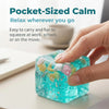 A hand squeezes a clear blue silicone fidget cube filled with small, colorful plastic spheres.