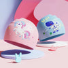 A pink silicone swimming cap shaped like a unicorn is displayed next to a blue space-themed cap.