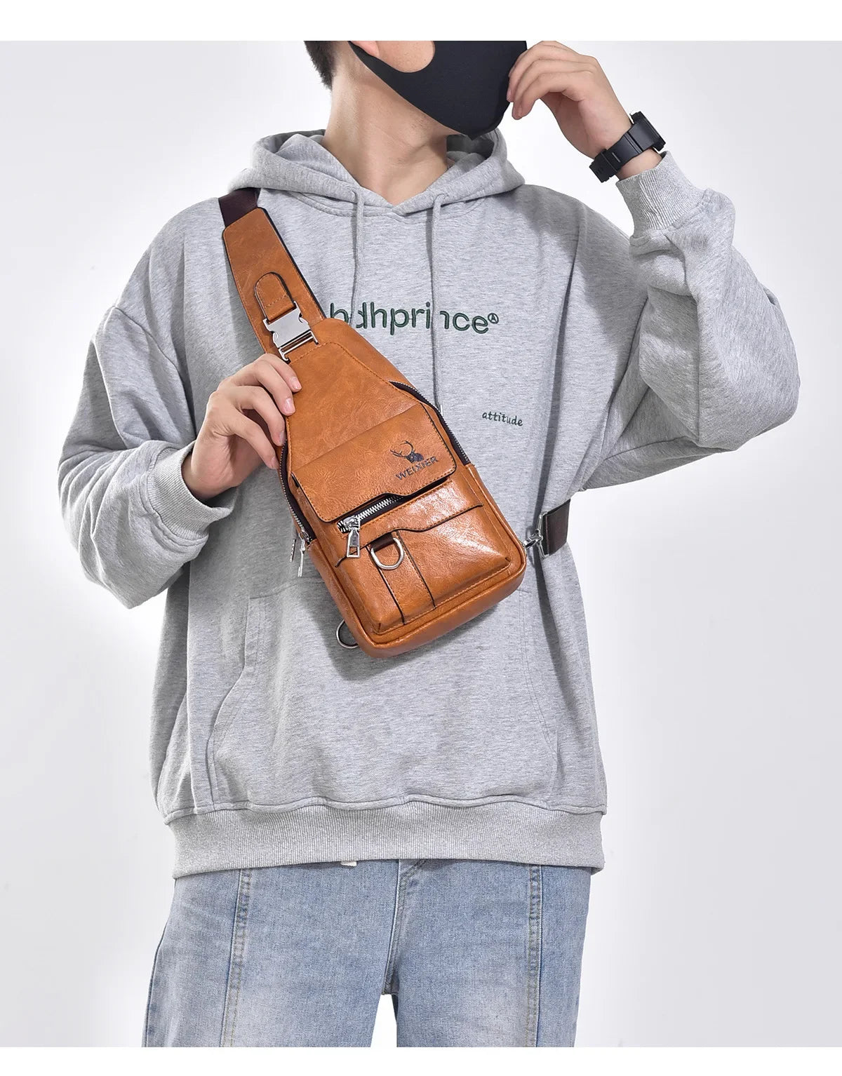 A man wearing a gray hoodie and jeans holds a brown leather crossbody bag.