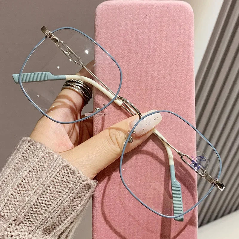 A hand holds a pair of thin blue-framed reading glasses resting on a pink surface.