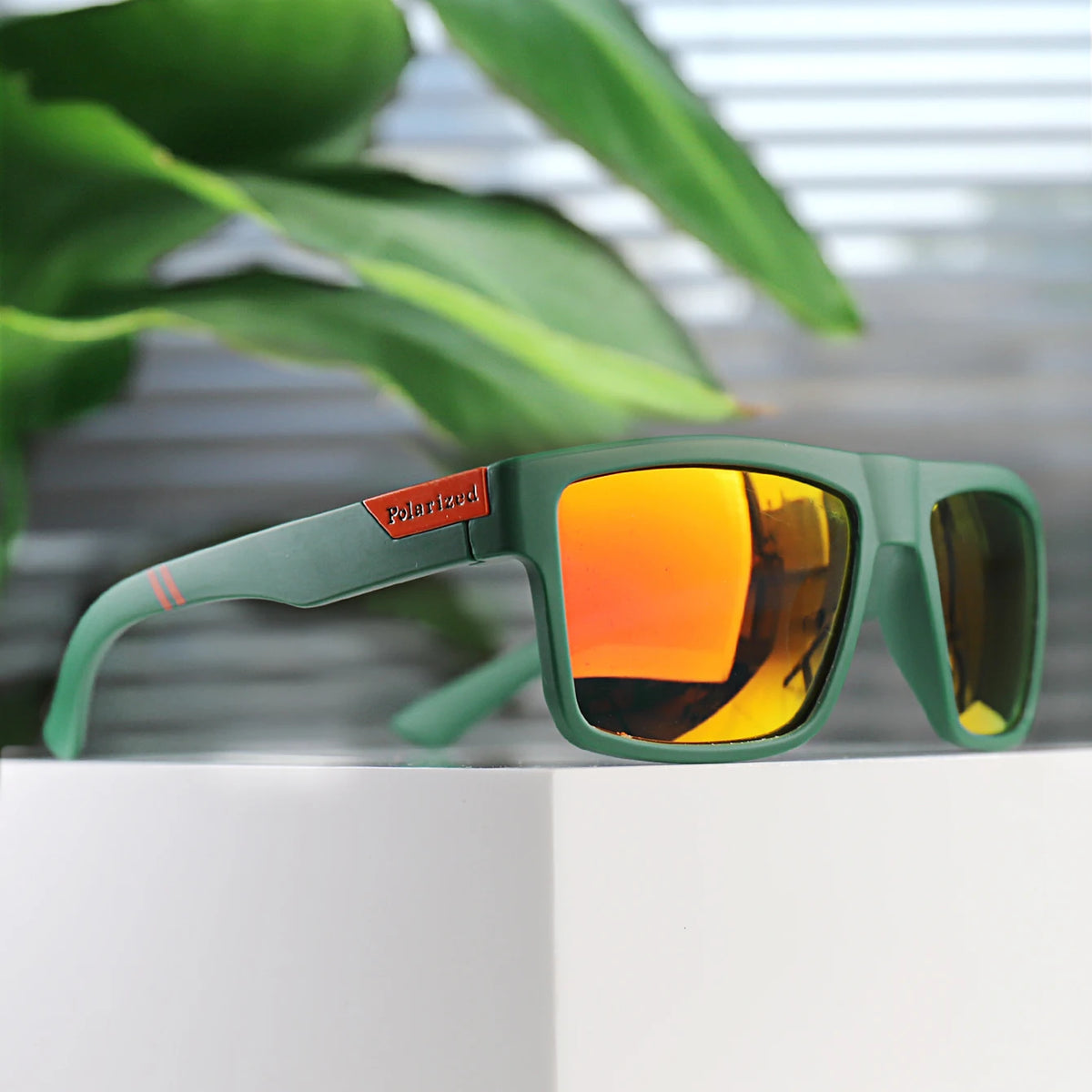 A pair of green polarized square sunglasses resting on a white surface with blurred green foliage in the background.