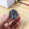 A hand holds a silver championship ring featuring a lightning bolt design and blue diamonds.