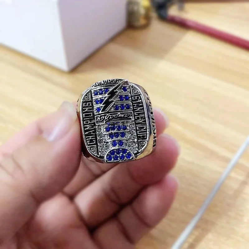 A hand holds a silver championship ring featuring a lightning bolt design and blue diamonds.