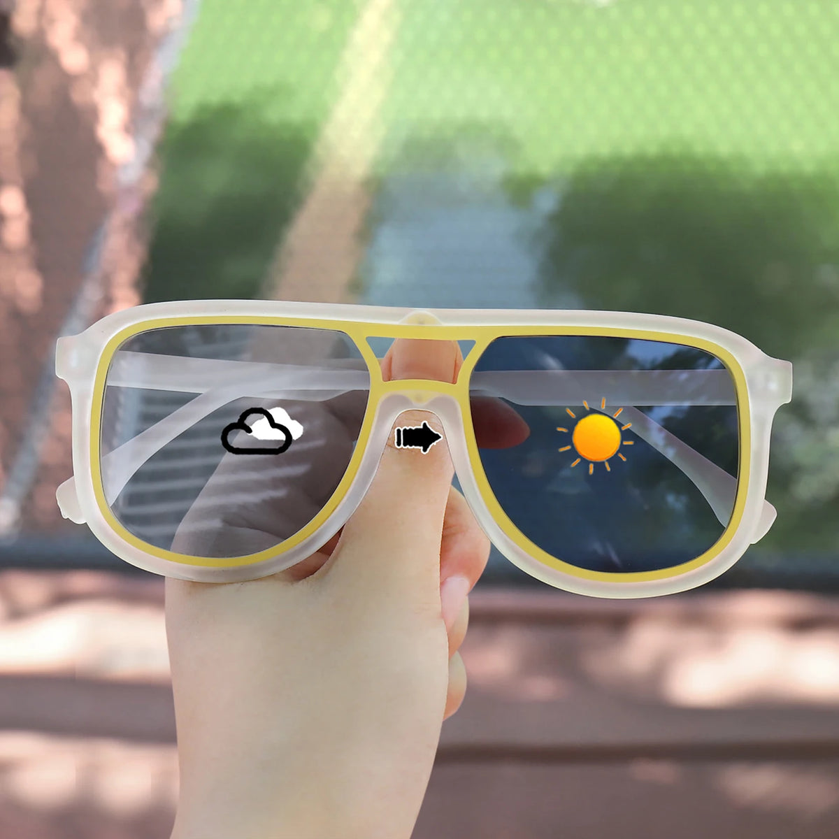 A hand holds a pair of square photochromic sunglasses with yellow frames and a lens showing a sun icon.