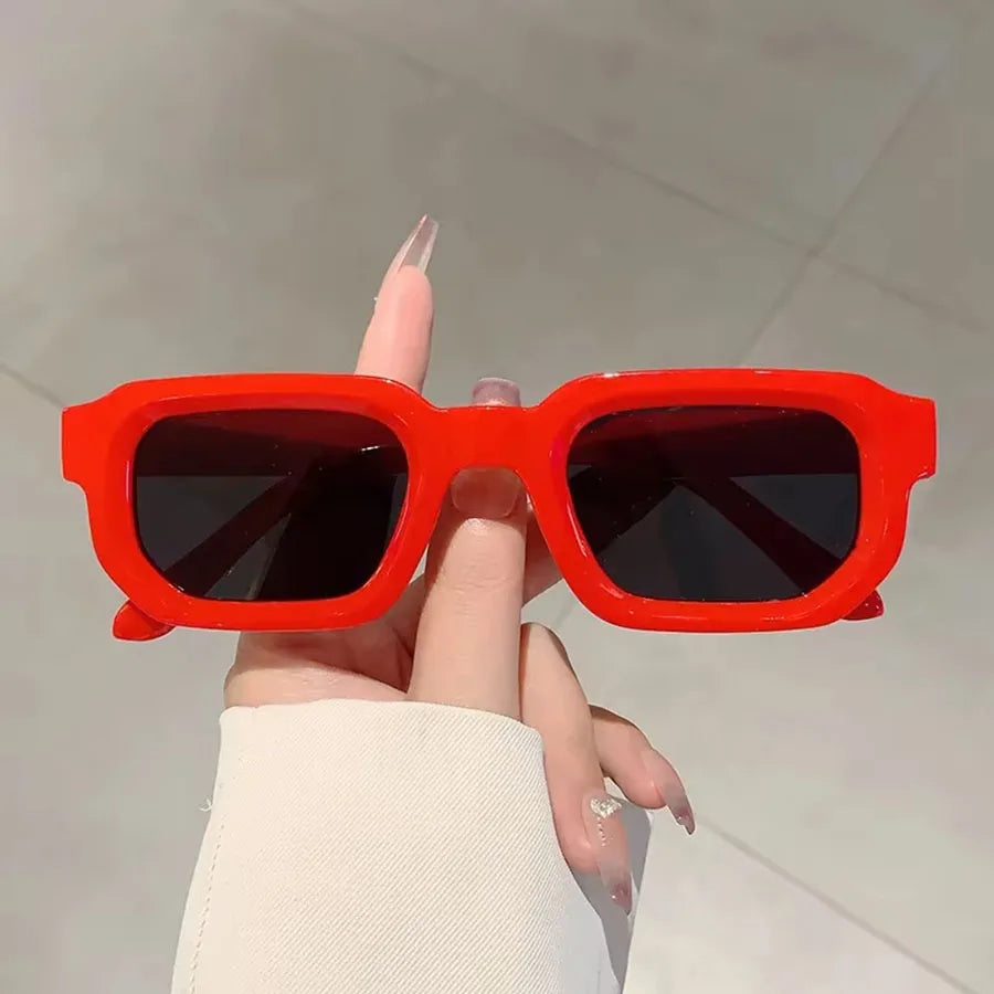 Une main tient des lunettes de soleil rectangulaires orange vif rétro sur un fond clair.