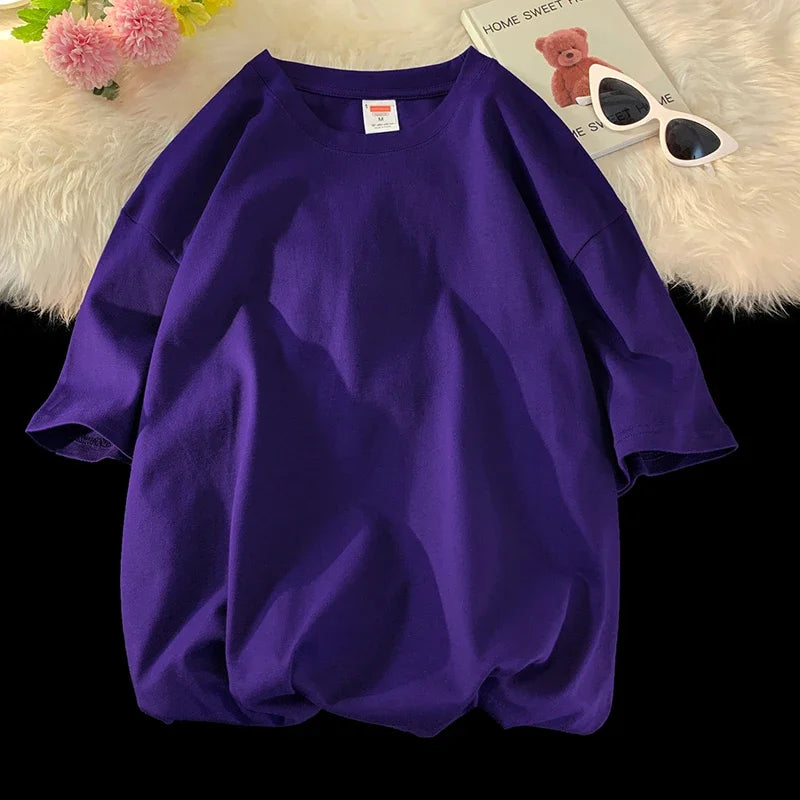 Lila, oversized Baumwoll-T-Shirt liegt auf weißem Fell neben Sonnenbrille und rosa Blume.