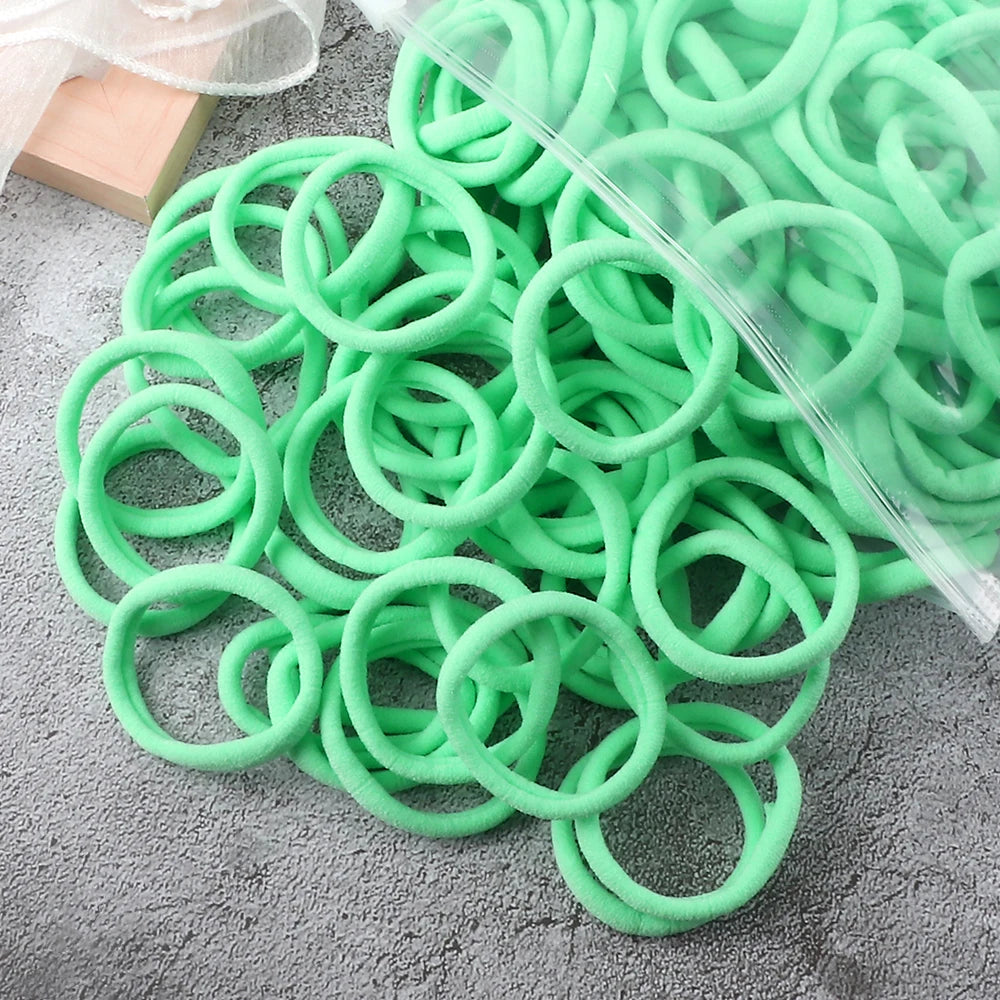 A pile of 50 pieces of mixed color elastic hair bands, some green and some clear, lying on a gray surface.