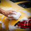A hand is applying a thick layer of white foam to the yellow paint of a car, demonstrating automotive gloss and protection.