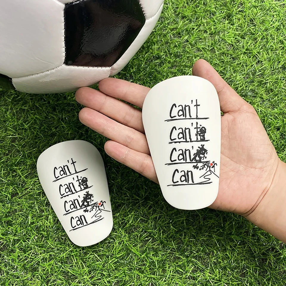 A hand holds a white mini wear-resistant shin guard with handwritten text on its surface, next to a soccer ball on green gras