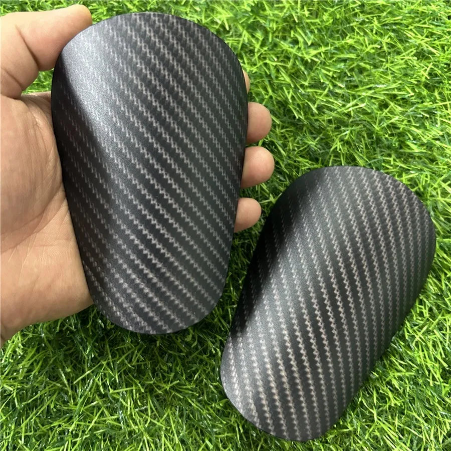 A pair of black carbon fiber-look soccer shin guards being held in a hand on green grass.