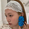 Primer plano de una mujer con una diadema de encaje floral blanca y un pendiente de flor de tela azul brillante.