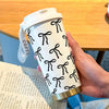 A hand holds a white double-wall insulated stainless steel travel mug with a black bow pattern on a wooden table.