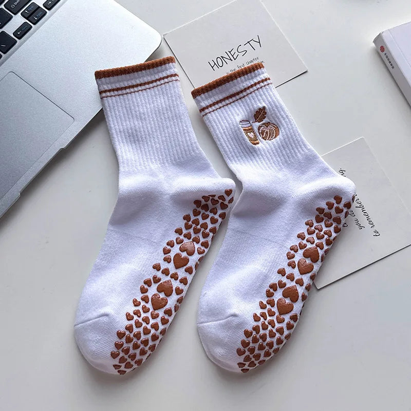 Paire de chaussettes de yoga blanches avec broderie de cœur marron et une petite feuille, posées sur une surface blanche.