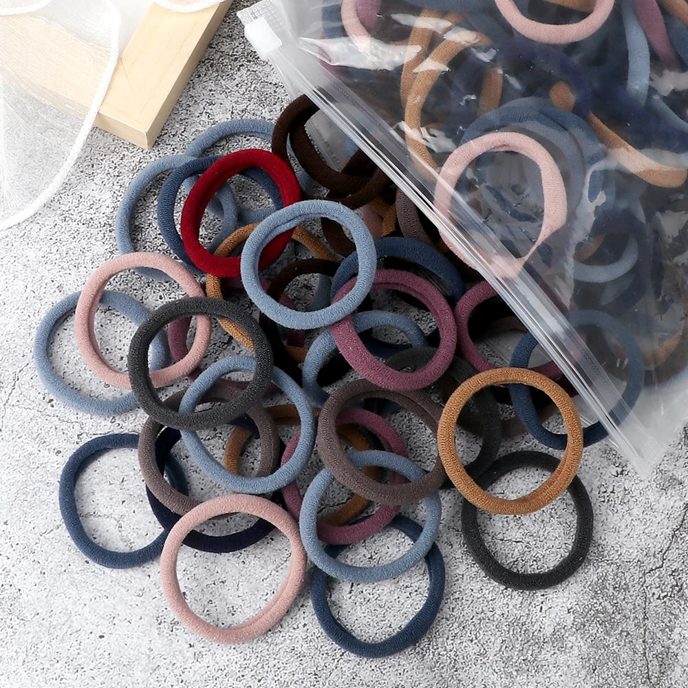A pile of 50 mixed color elastic hair bands in various shades of gray, brown, and pink, with some packaged in clear plastic.