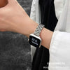 A close-up of a black smartwatch with a stainless steel band is worn on a wrist next to a white garment.