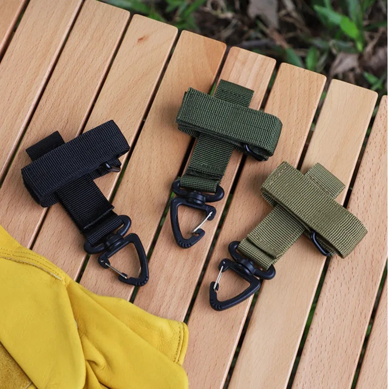 Two black and olive green tactical nylon glove hooks resting on a wooden surface next to a yellow glove.