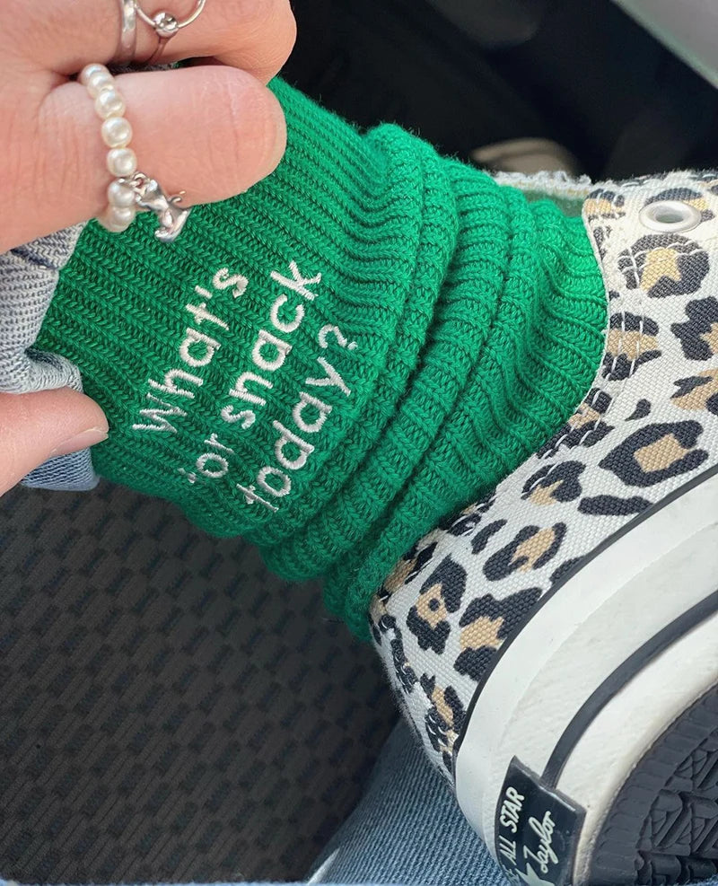 A hand is holding a bright green knitted sock with white embroidery that reads What's snack or snack today on a leopard print