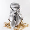 A white satin square scarf with black polka dots tied in a bow, displayed on light blonde hair.