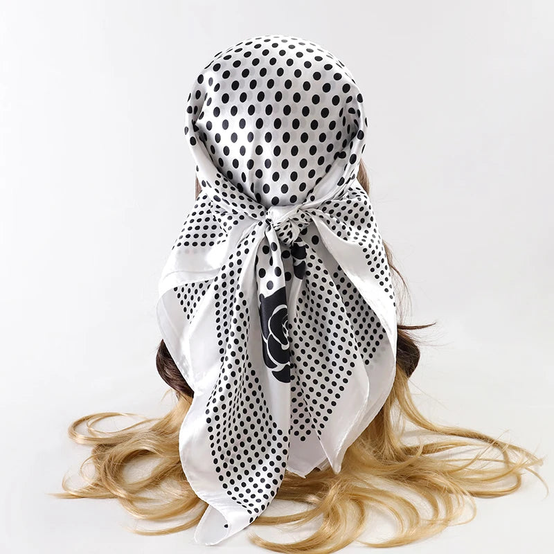 A white satin square scarf with black polka dots tied in a bow, displayed on light blonde hair.